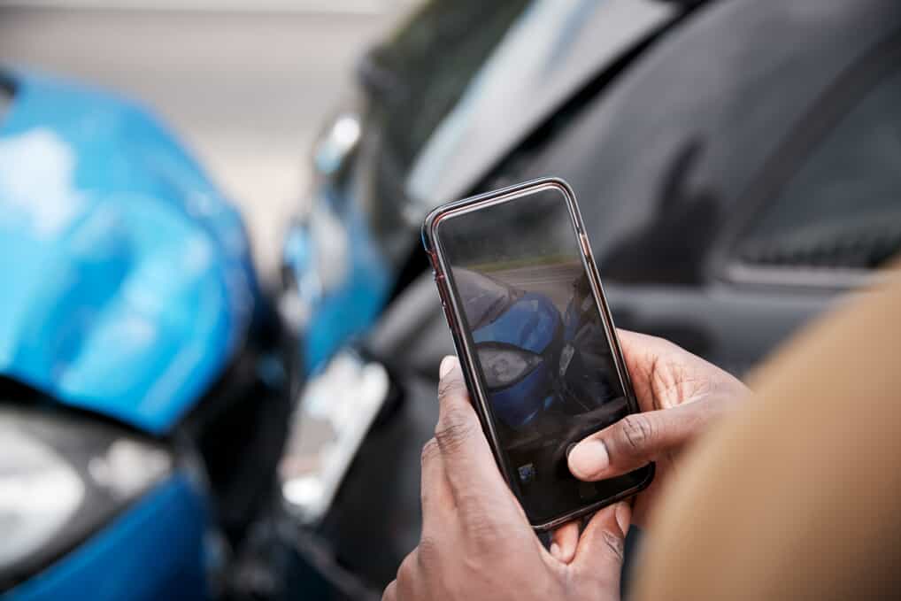 Man's hands holding a smartphone as he takes a photo of the damage to the collided vehicles